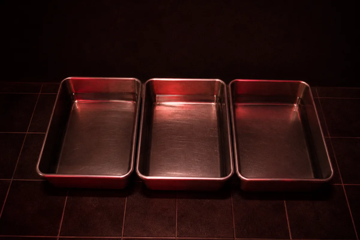 Bacs de développement en inox posés au sol d&rsquo;une chambre noire, remplis d&rsquo;un liquide clair sous lumière rouge