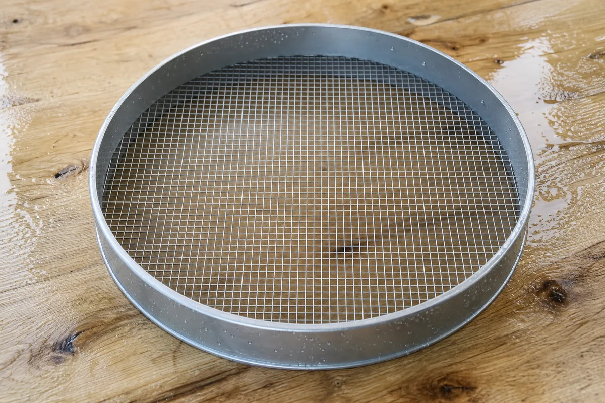 Passoire en inox posée sur une planche de bois humide, gouttes d’eau retenues dans les perforations